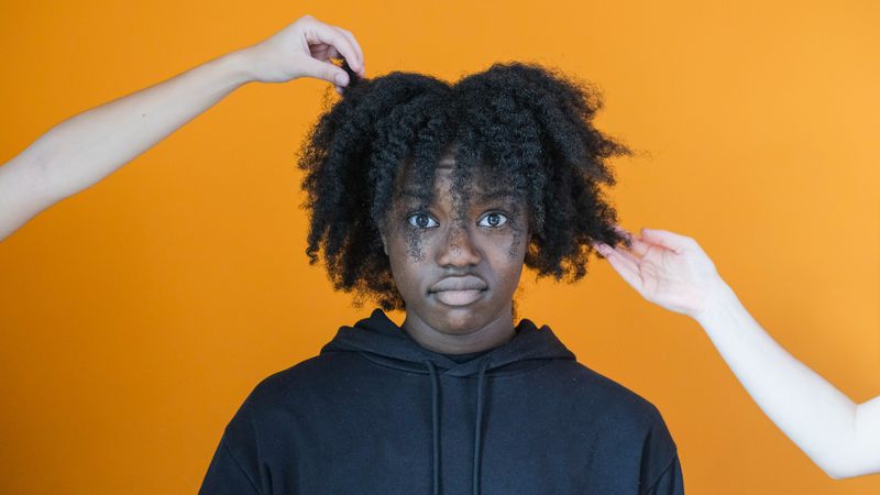 Photo d'une jeune femme noire aux long cheveux bouclés crépus que deux mains blanches touchent sans demander de consentement